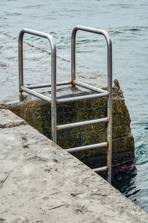 Grey concrete pier with metal ladder at blue sea water with small waves on an overcast summer dayの写真素材