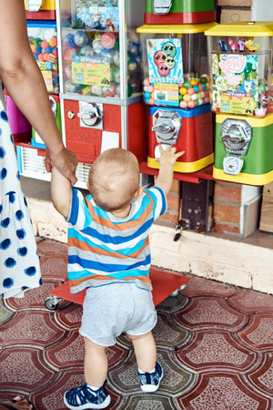 Gelendzhik, Russia - August 09 2021: Excited toddler boy wants to win toys in machine holding mother hand at sea resort. Fair-haired kid tries to force parent to buy and win toyのeditorial素材
