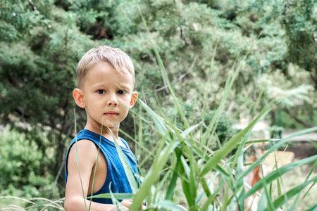 Cute blond boy stands near lush plant with long green leaves looking in camera. Curious preschooler enjoys nature at summer resort closeupの写真素材
