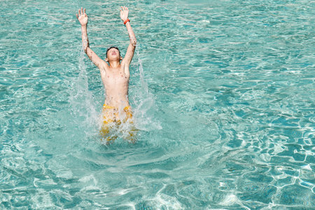 Happy boy jumps out of the water in swimming pool with raising hands while posing for photo in water park on sunny dayの写真素材