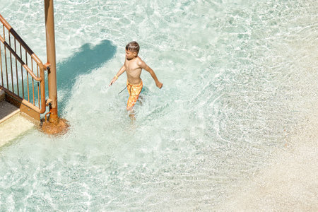 Schoolboy goes out of swimming pool with clear blue water on sunny day. Blond boy enjoys fun activities in aqua park on summer vacationの写真素材