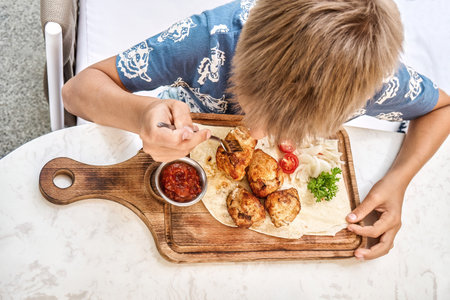 Blond boy eats grilled meat dish sitting at table in restaurant close upper view. Preteen child enjoys nutritious meal in cafeteriaの写真素材