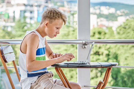 Preteen boy sitting on hotel balcony and surfing the internet on grey laptop. Schoolboy enjoys spending summer holidays and playing gamesの写真素材