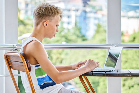 Preteen boy sitting on hotel balcony and surfing the internet on grey laptop. Schoolboy enjoys spending summer holidays and playing gamesの写真素材