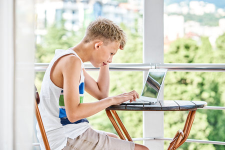 Preteen boy sitting on hotel balcony and surfing the internet on grey laptop. Schoolboy enjoys spending summer holidays and playing gamesの写真素材