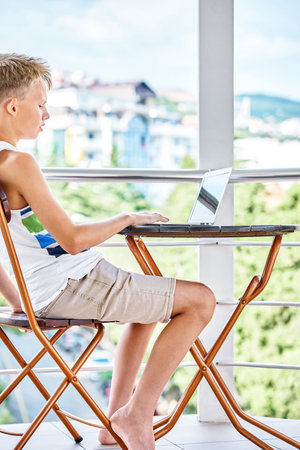 Preteen boy sitting on hotel balcony and surfing the internet on grey laptop. Schoolboy enjoys spending summer holidays and playing gamesの写真素材