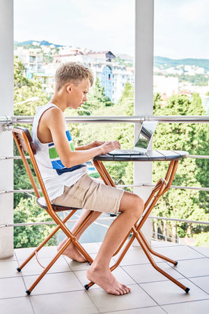 Preteen boy sitting on hotel balcony and surfing the internet on grey laptop. Schoolboy enjoys spending summer holidays and playing gamesの写真素材