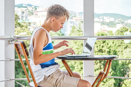Preteen boy sitting on hotel balcony and surfing the internet on grey laptop. Schoolboy enjoys spending summer holidays and playing gamesの写真素材