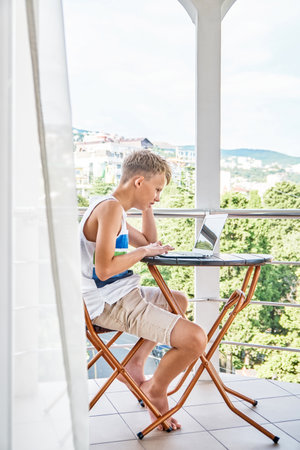 Preteen boy sitting on hotel balcony and surfing the internet on grey laptop. Schoolboy enjoys spending summer holidays and playing gamesの写真素材