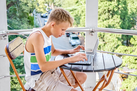 Preteen boy sitting on hotel balcony and typing messages to friends on grey laptop. Schoolboy enjoys spending summer holidays and playing gamesの写真素材