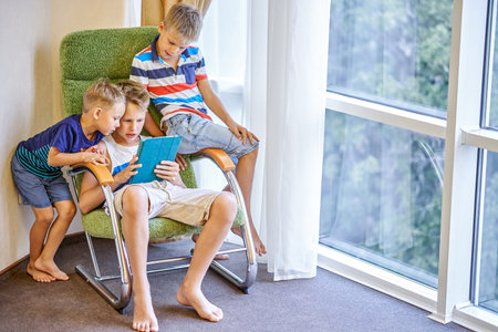 Boys siblings spend time playing interesting games on tablet together. Younger brothers watching preteen schoolboy playing game in hotel roomの写真素材