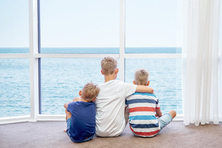 Eldest boy hugs younger brothers sitting on floor and looking at azure sea from panoramic window. Siblings enjoy spending summer holidays togetherの写真素材
