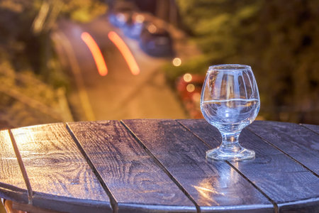 Empty wineglass on dark wooden table on hotel balcony against evening resort town. Glass illuminated by spotlight against road with carsの写真素材