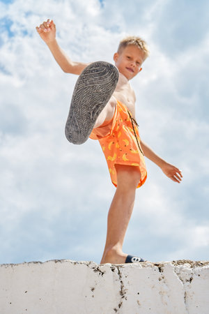 Junior schoolboy with delighted and smiling expression on high pier against blue sky with clouds. Boy raises foot upward enjoying spending summer holidays at seaside. Selective focusの写真素材