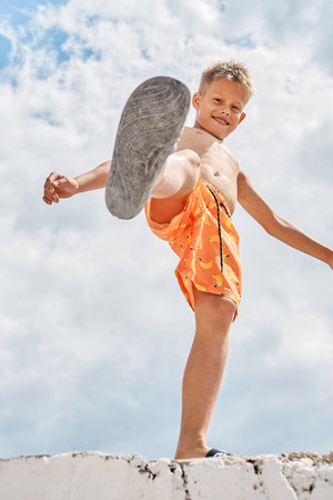 Junior schoolboy with delighted and smiling expression on high pier against blue sky with clouds. Boy raises foot upward enjoying spending summer holidays at seasideの写真素材