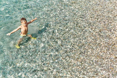 Preschooler boy enjoys exploring sea water on sunlit beach. Blond kid walking on pebble bottom of sunny sea at beach resort upper viewの写真素材
