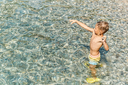 Preschooler boy in bright colorful shorts standing in sea water and throws stones. Child enjoy summer holidays on seaの写真素材