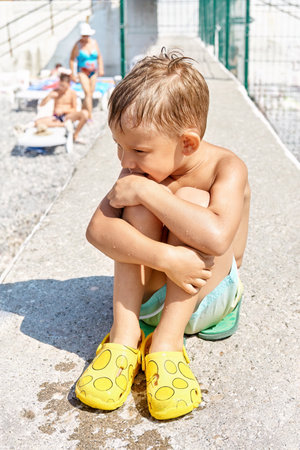 Preschooler boy sitting on sunny pier relaxes from swimming in sea. Kid with sad expression wants to spend more time in sunlit waterの写真素材
