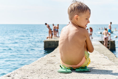 Preschooler boy sitting on sunny pier relaxing from sea activities. Child enjoys spending summer holidays with family at sea resortの写真素材