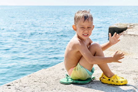 Preschooler boy sitting on sunny pier relaxes from swimming in sea. Kid with sad expression wants to spend more time in sunlit waterの写真素材