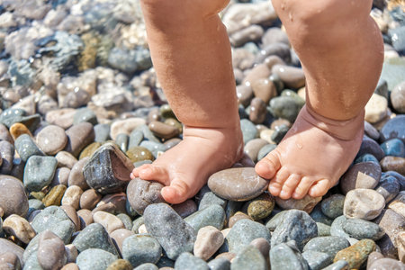 Infant boy standing on pebbles with wet feet on sunny beach. Child casting shadow explores pebbles on sunlit sea beach at seaside resortの写真素材