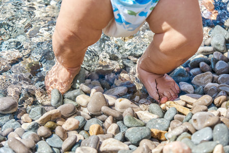 Infant boy standing on pebbles with wet feet on sunny beach. Child casting shadow explores pebbles on sunlit sea beach at seaside resortの写真素材