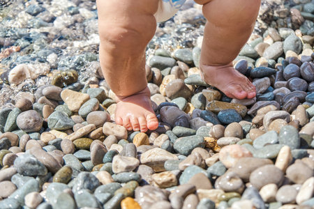 Infant boy standing on pebbles with wet feet on sunny beach. Child casting shadow explores pebbles on sunlit sea beach at seaside resortの写真素材