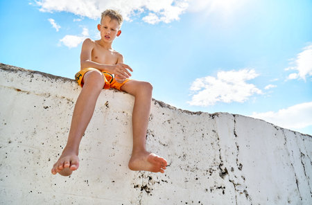 Preteen boy sits on sunny pier watching seaside against blue sky. Blond schoolboy spending summer vacation at sunny seaside resortの写真素材