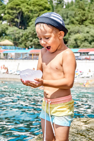 Preschooler boy standing on stone pier holds transparent jellyfish. Boy with amused and surprised expression looks at jellyfish in handsの写真素材
