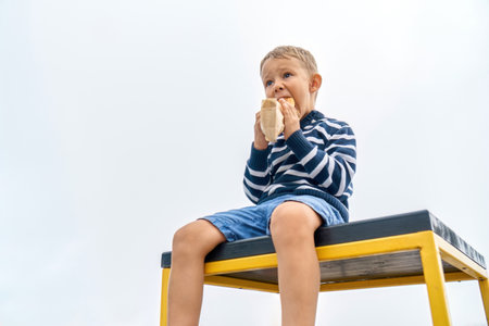 Hungry little boy with hood eats sandwich sitting on table on misty day in mountain park. Cute child bites snack resting in foggy highland. Kid has dinner outdoorsの写真素材