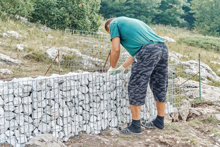 Gelendzhik, Russia - August 17, 2021: Gabion wall building process. Builder puts rocks in metal mesh to make gabion wall in mountain park. Man fills net basket with stones building traditional fenceのeditorial素材