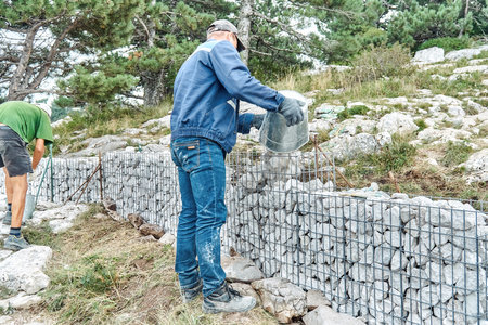 Gelendzhik, Russia - August 17, 2021: Gabion building process. Builder puts rocks in metal mesh to make gabion in mountain park. Man fills net basket with stones building traditional fenceのeditorial素材