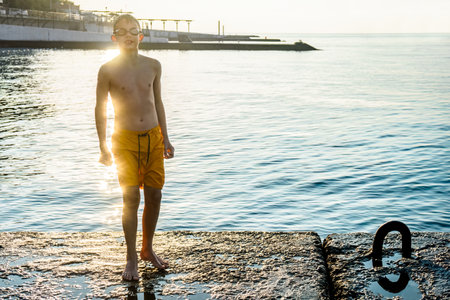Boy with swim goggles and wet shorts stands on stone pier in cool morning. Sportive kid rests after refreshing swimming in cold sea waterの写真素材