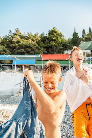 Laughing brothers stand together on the beach at sea resort. Joyful boys with towel and clothes rest after swimming. Happy children on vacationの写真素材