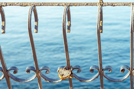 Heart shaped padlock on old metal fence by transparent sea bay closeup. Strong love symbol on grid against ocean water on sunny day. Cute artefactの写真素材