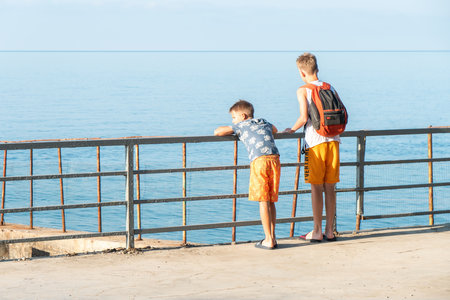 Little boys observe sea standing on ground at summer resort. Children lean on handrail looking at calm beach in morning. Kids on vacationの写真素材