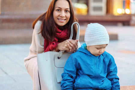 Smiling mother sits near little son playing on city street. Woman and adorable baby walking on town square portrait. Lady with offspringの写真素材