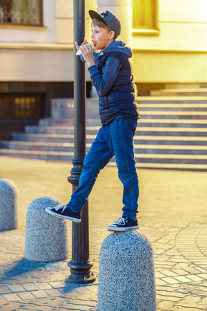 Playful boy eats ice cream standing on stone pillar in evening city. Kid licks sundae in cone having fun on the street. Urban lifestyle and street foodの写真素材