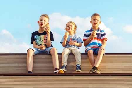 Little boys brothers eats tasty chocolate ice cream sitting on wooden bench watching movie on huge screen against sky with clouds in parkの写真素材