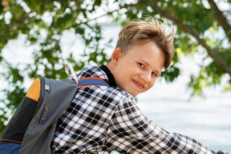 Portrait of smiling boy with blond hair in plaid shirt with backpack sitting on bicycle, on blurred...
