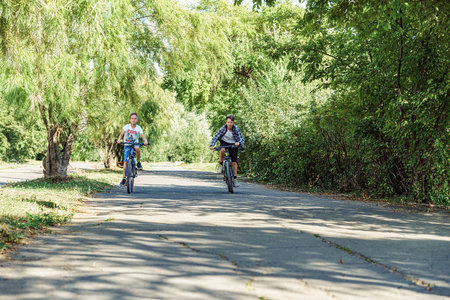 Two joyful boys riding bicycles on bicycle lane in park with lush foliage on warm and sunny summer day, enjoying freedom and beauty of outdoorsの写真素材