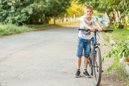 Tired boy in a t-shirt and denim shorts leans on his bicycle after a long bike ride in the park, showing exhaustion and satisfactionの写真素材