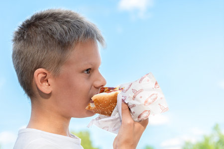 Hungry boy in white T-shirt savoring a hot dog outdoors on a sunny day, taking a satisfying bite with a serene blue sky and fluffy white cloudの写真素材