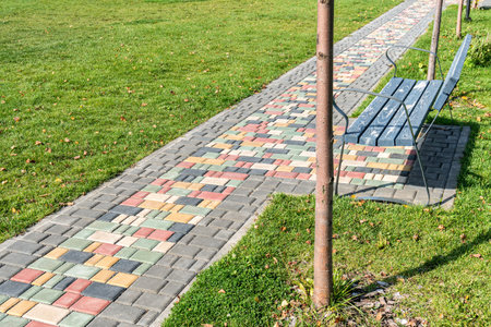 Colorful paved road across green lawn with empty bench on sunny day. Pathway and seat in summer park. Tiled pathway across public gardenの写真素材