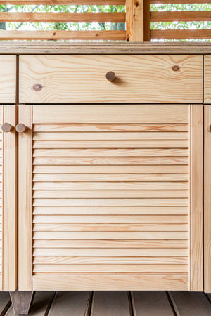 Wooden cabinets of outdoor kitchen with louvered doors, drawers, wooden handles and a white plywood countertop, stand on a wooden terrace close-up viewの写真素材
