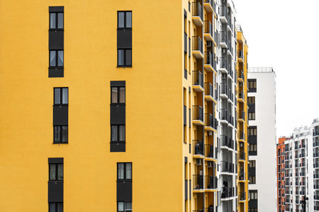 Modern apartment buildings with yellow and white facades, large windows, and balconies with metal railings, against a cloudy sky, urban livingの写真素材