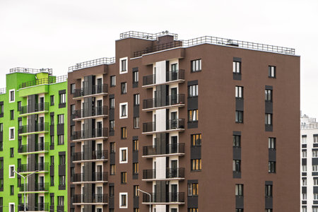 Modern apartment buildings with green and brown facades, large windows, and balconies with metal railings, standing against a cloudy sky, urban livingの写真素材
