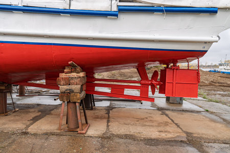Dry-docked boat with a red and white hull, showing its rudder system and propeller, supported by...