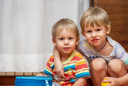 Two young boys sitting close together indoors, one arm around the other, with a curtain and wooden background. Concept of childhood bondの写真素材