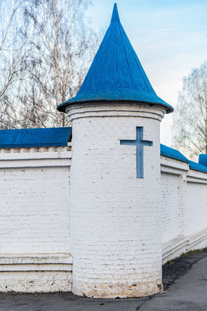 White brick monastery wall with blue-roofed towers and blue cross, with bare trees and clear sky, symbolizing religionの写真素材